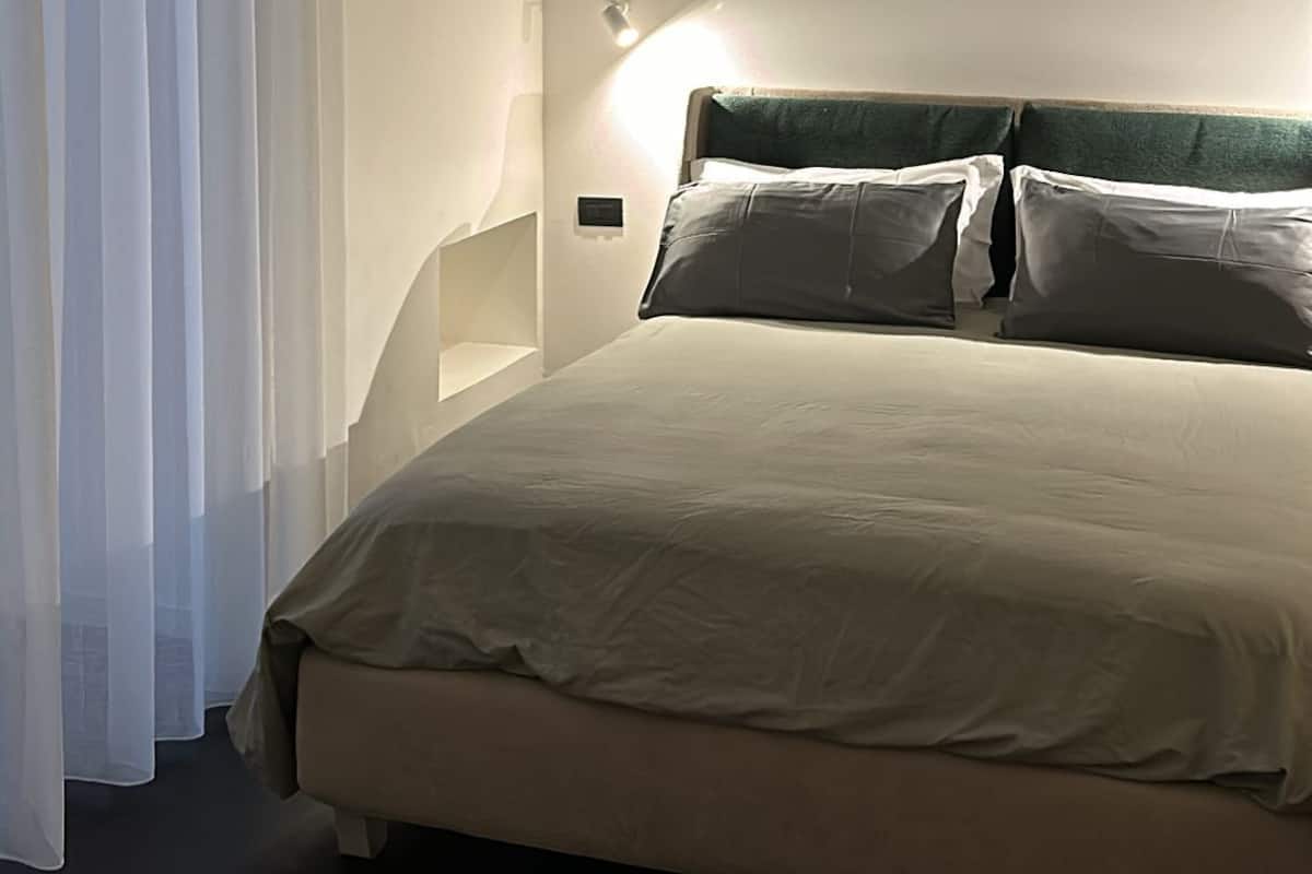 A bedroom with a bed, white curtains, a wall-mounted light, and a ceiling vent.