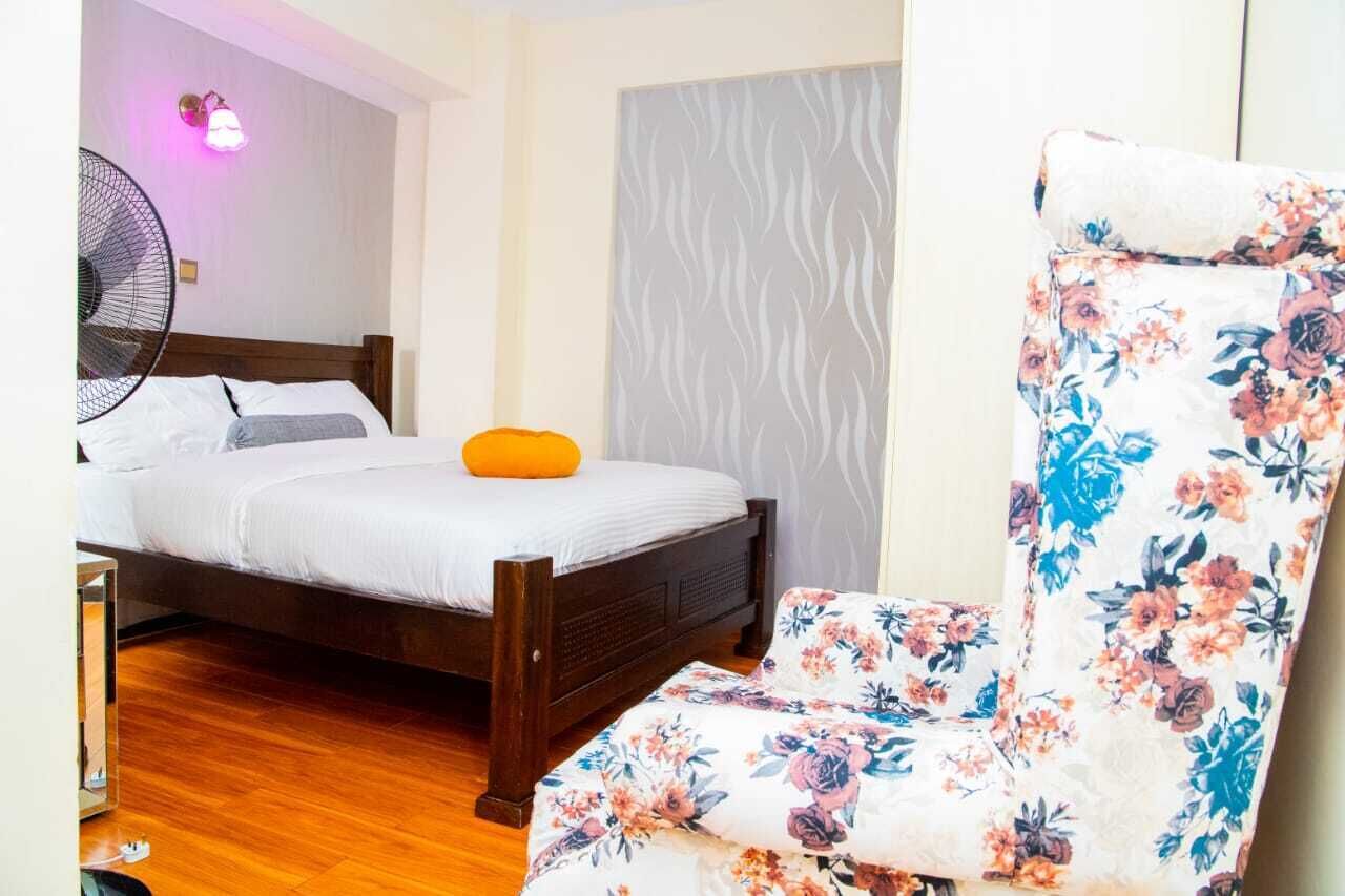 A bedroom with a wooden bed, a large floral armchair, a standing fan, and a wall-mounted light fixture.