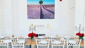 Un comedor con sillas blancas, una mesa dispuesta para seis personas y un cuadro de un campo con un árbol en la pared.