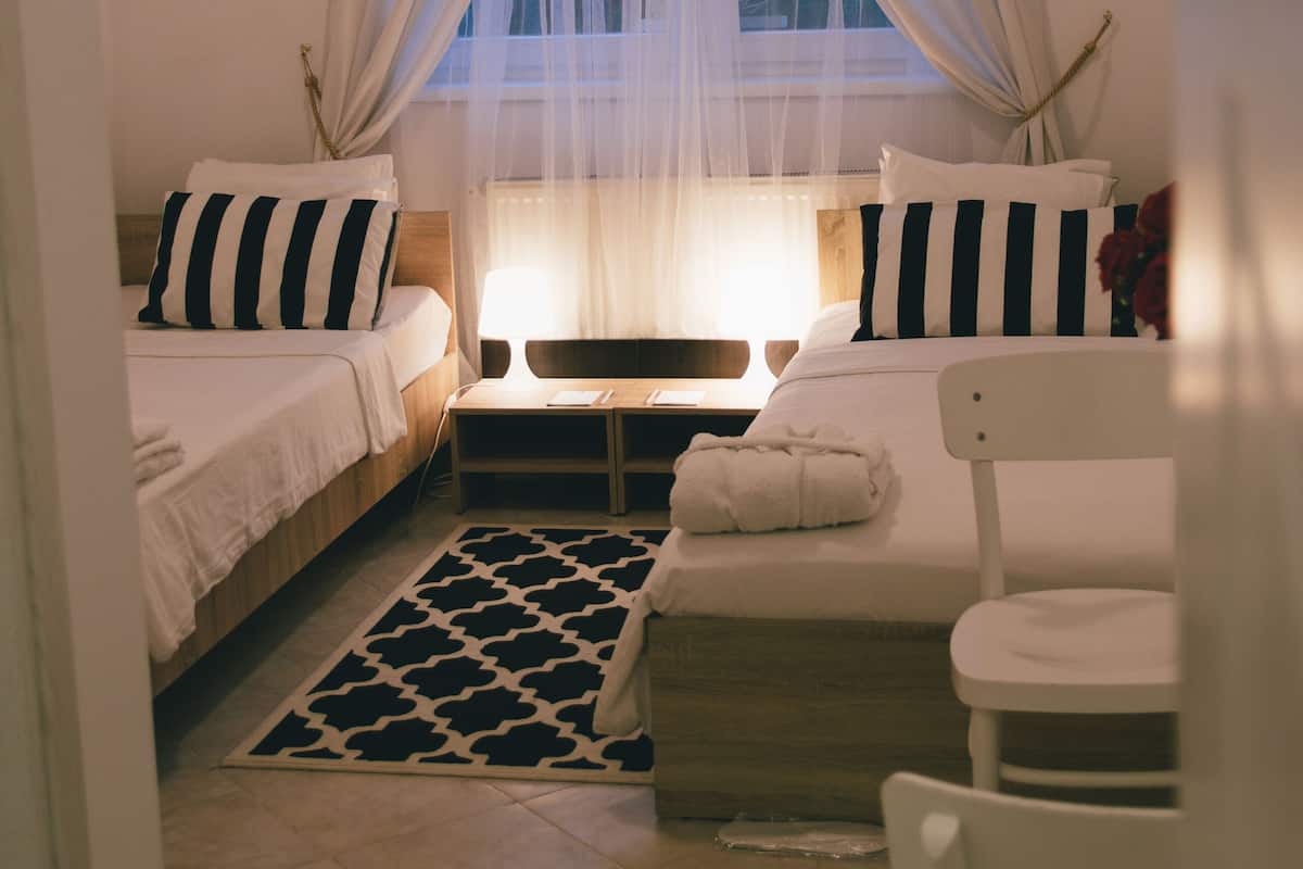 A bedroom with two beds, a bedside table, a chair, a window with sheer curtains, and a framed sign on the wall.