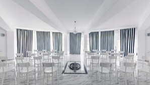 Una habitación amplia con piso de mármol, sillas blancas dispuestas en filas y cortinas a rayas negras y blancas.