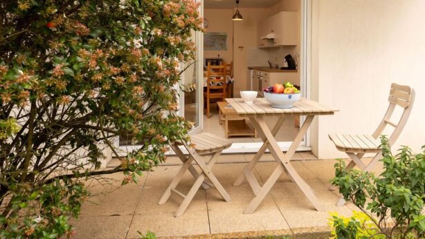 A wooden table with chairs and a bowl of fruit in a patio area with a view of a kitchen and dining area. - L'air du Large by Interhome (Saint Malo)