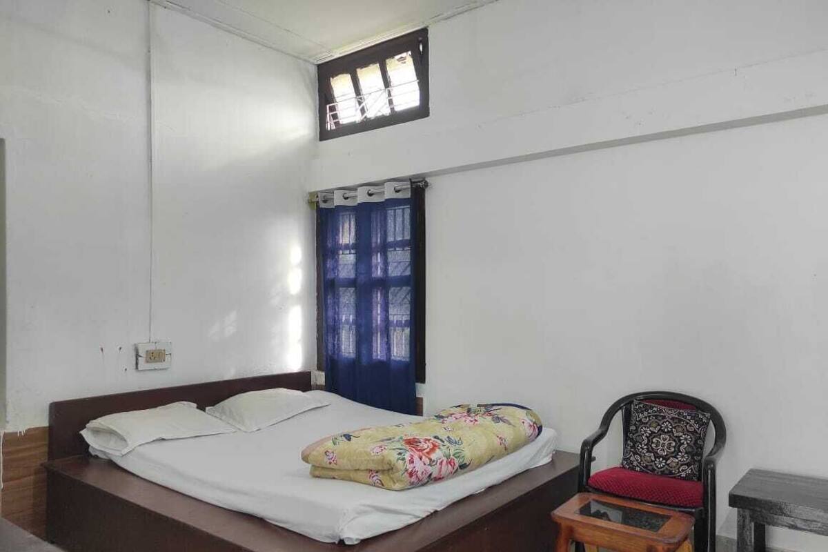 A bedroom with a single bed, a small table, a chair, a window with curtains, and a power outlet on the wall.