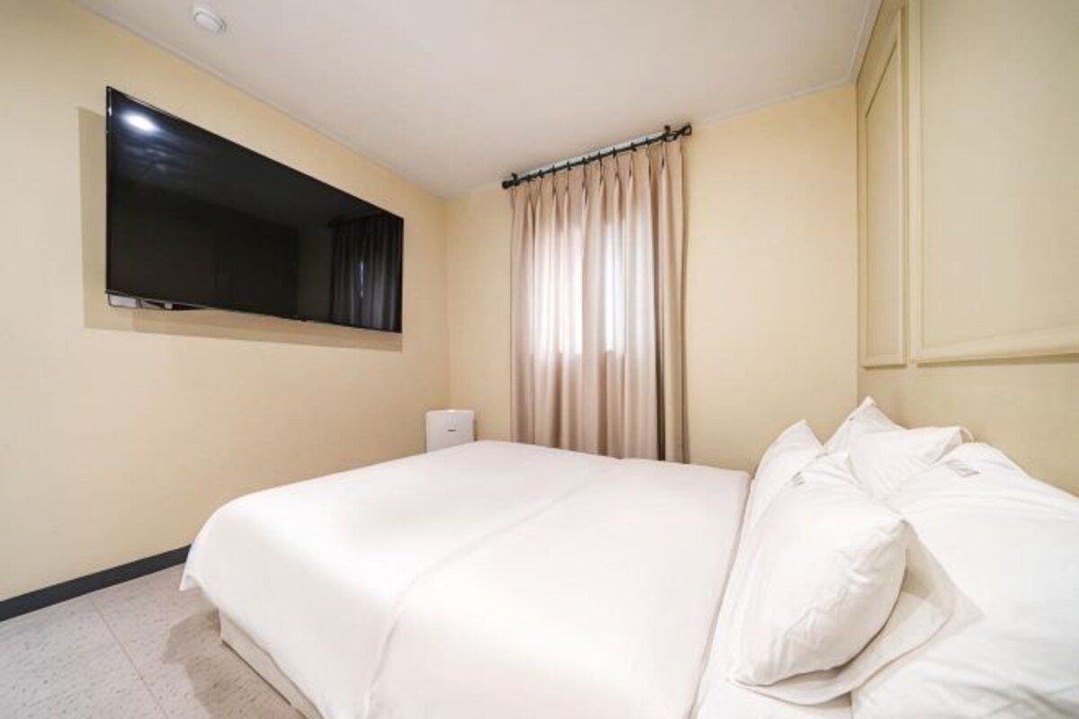 Hotellihuone, jossa on suuri sänky, seinälle asennettu taulutelevisio, ikkuna verhoilla ja yöpöytä.