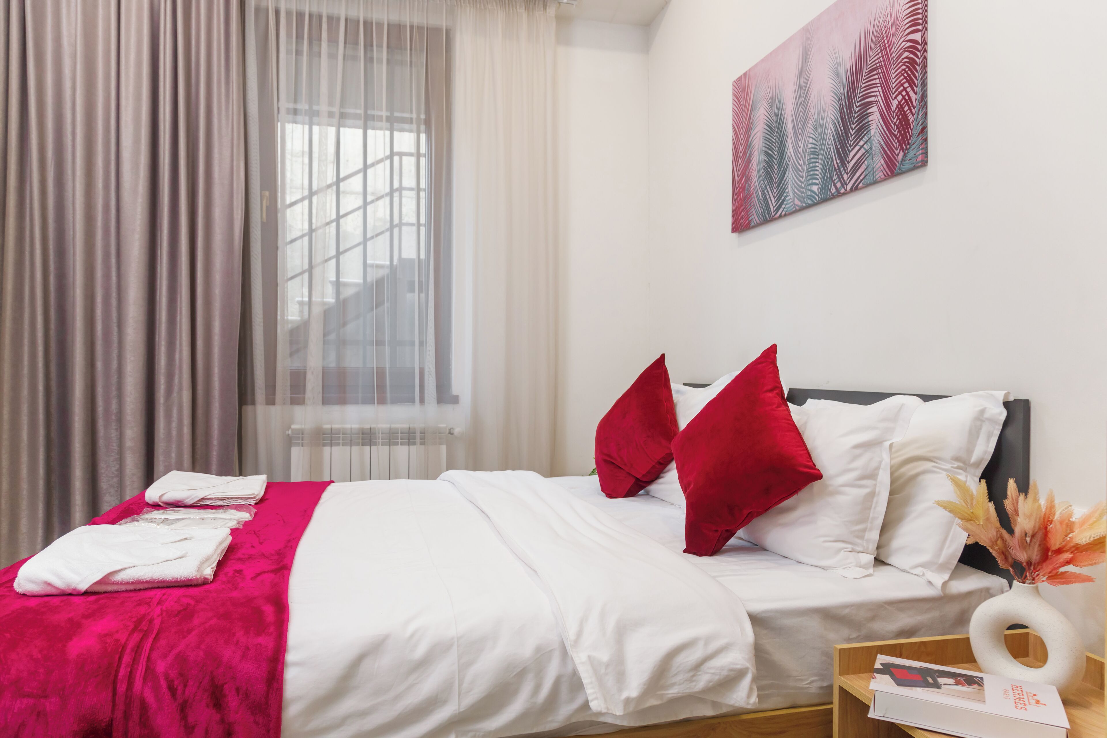 A neatly made bed with white and red pillows, a pink blanket, and a wooden side table with a book and a vase.