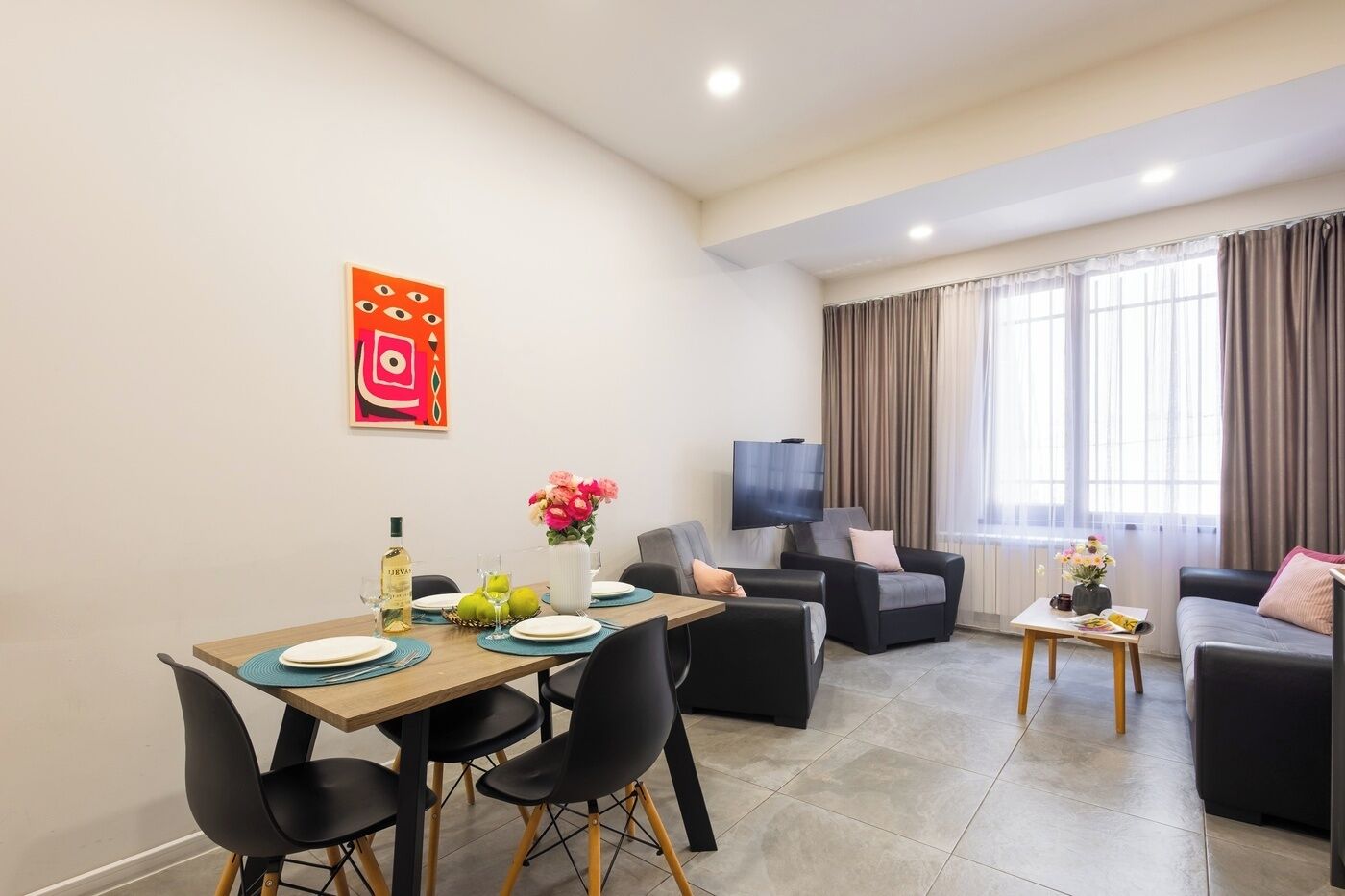 A modern living room with a dining area, a flat-screen TV, and a large window with curtains.