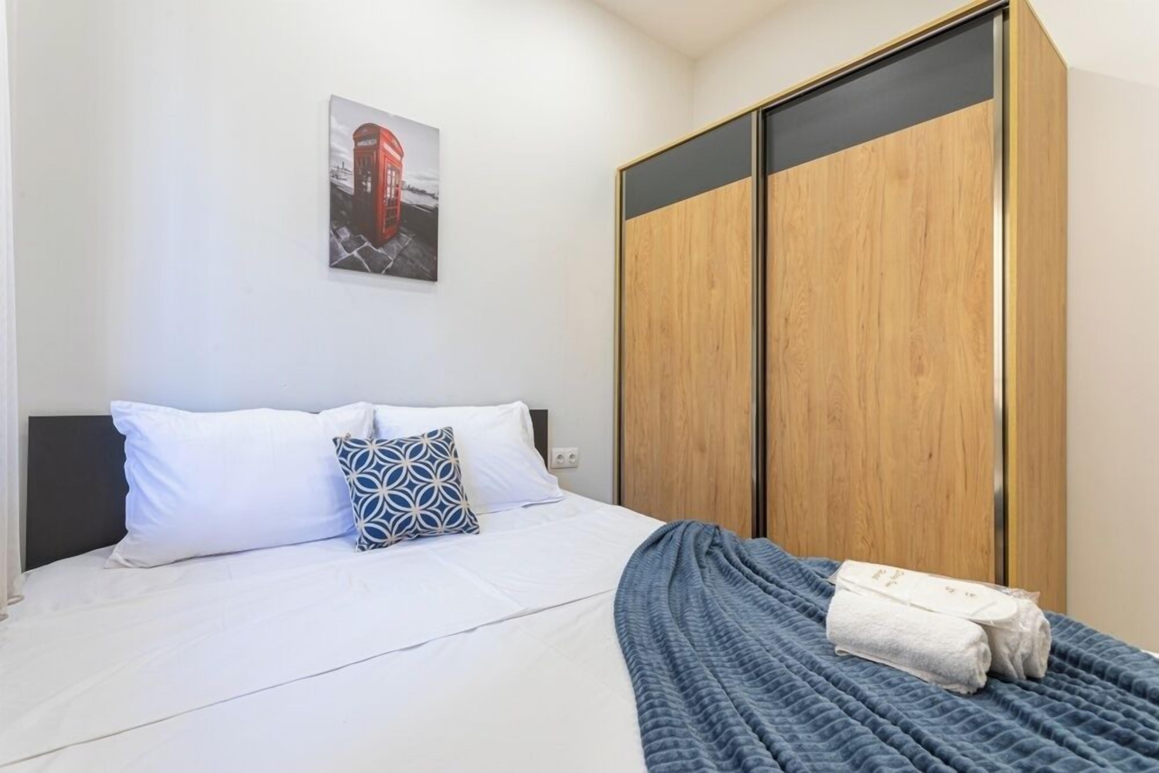 A neatly made bed with white bedding and a blue blanket, a wooden wardrobe with a black panel, and a picture of a red telephone booth on the wall.