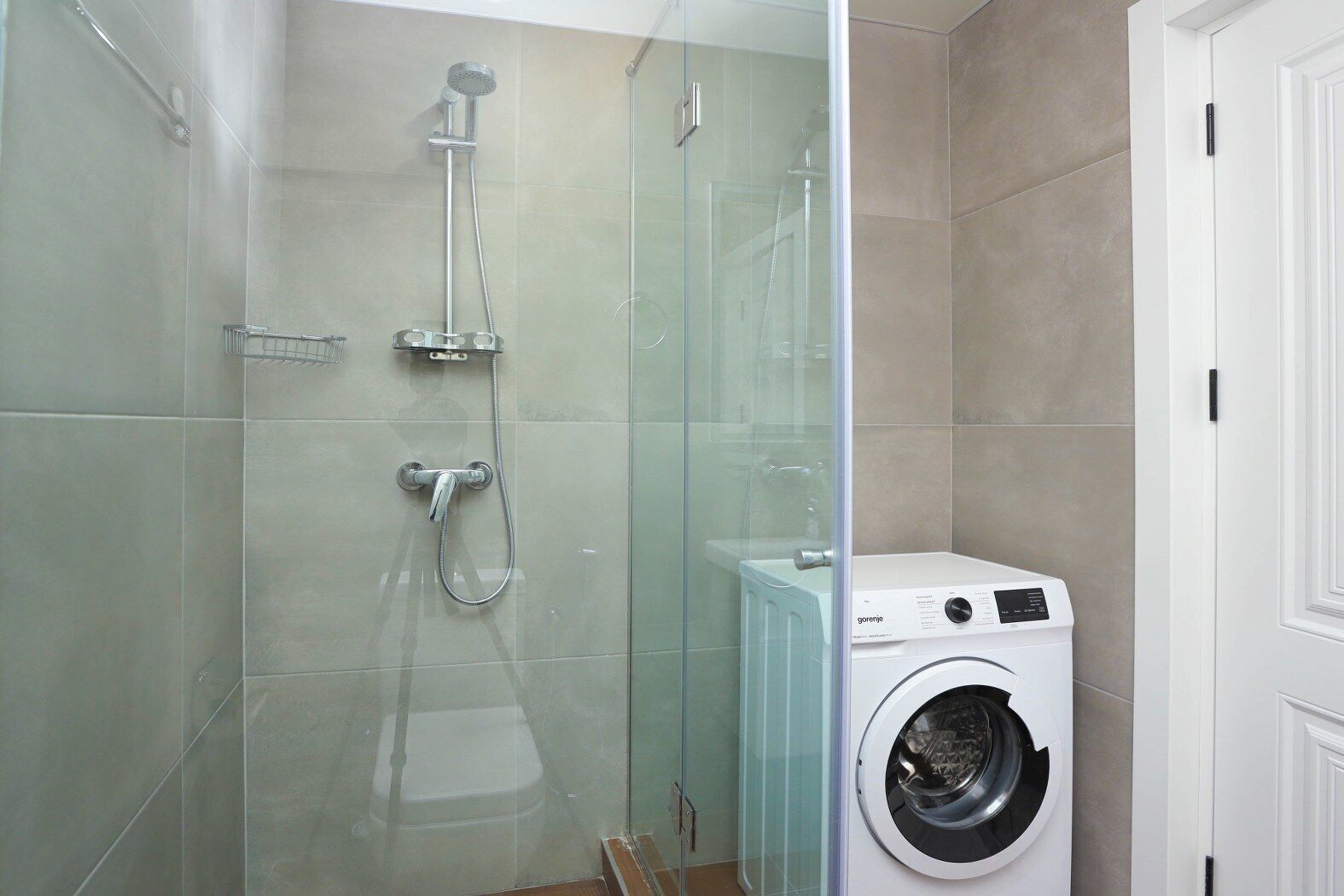 Un baño con ducha envuelta en vidrio, lavadora blanca y una puerta blanca.