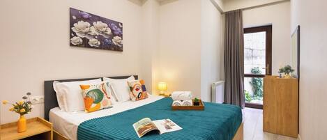 A hotel room with a bed, a nightstand, a bedside table, a window with curtains, and a view of greenery outside.