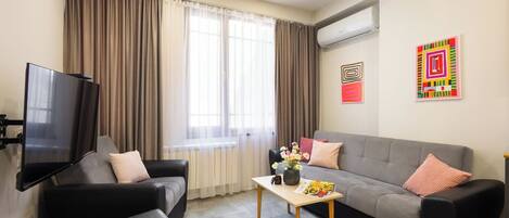 A modern living room with a flat-screen TV, two sofas, a coffee table, and a window with curtains.