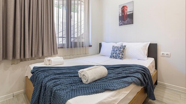 Un lit bien fait, avec une couverture bleue et une serviette enroulée, une fenêtre avec des rideaux légers et un tableau accroché au mur.