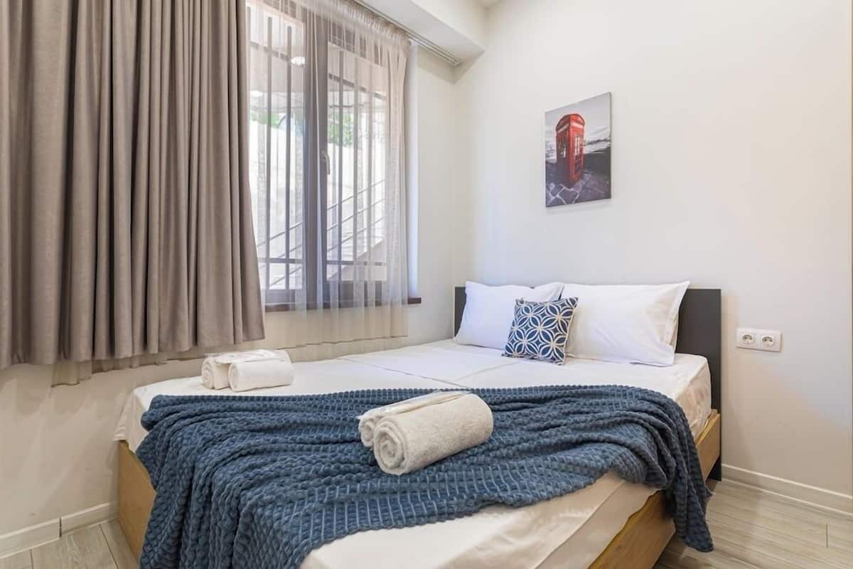 Un lit bien fait, avec une couverture bleue et une serviette enroulée, une fenêtre avec des rideaux légers et un tableau accroché au mur.