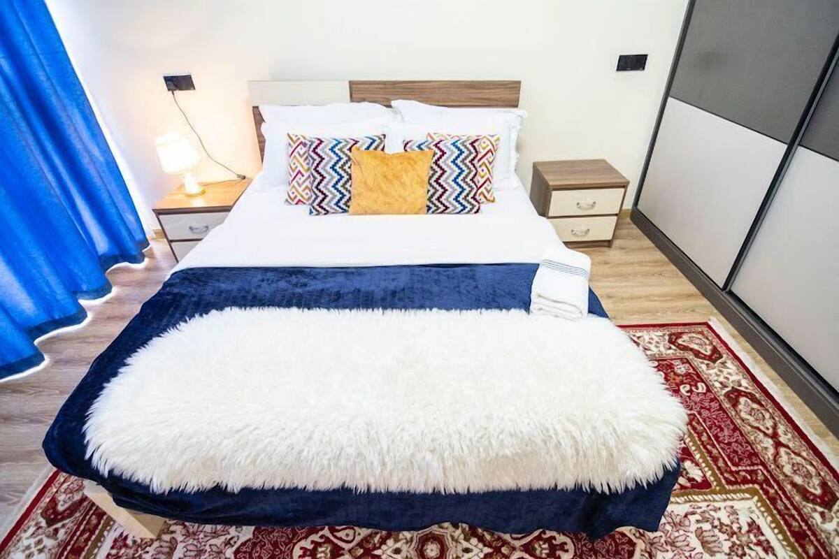Ein Bett mit einem weißen, flauschigen Kissen, einer blauen Decke und einem gemusterten Kopfkissen. Ein Nachttisch mit einer Lampe, einer Holoszene und einem roten Teppich mit Muster.
