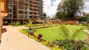 A modern building with multiple balconies, a well-maintained garden with various plants, and a clear sky. - Lux Suites Urban Oasis Apartments (Nairobi)