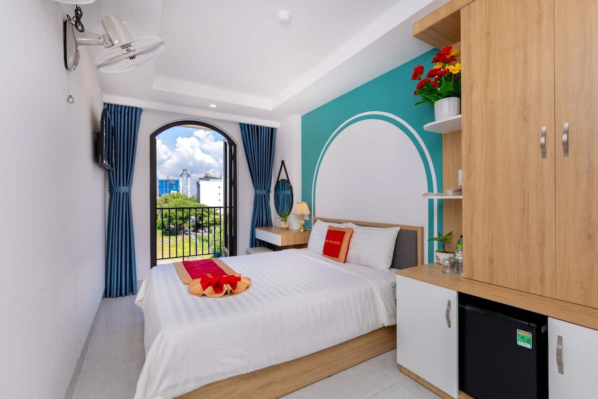 Habitación de hotel con cama, un pequeño escritorio, armario, balcón con cortinas y vista a la ciudad.