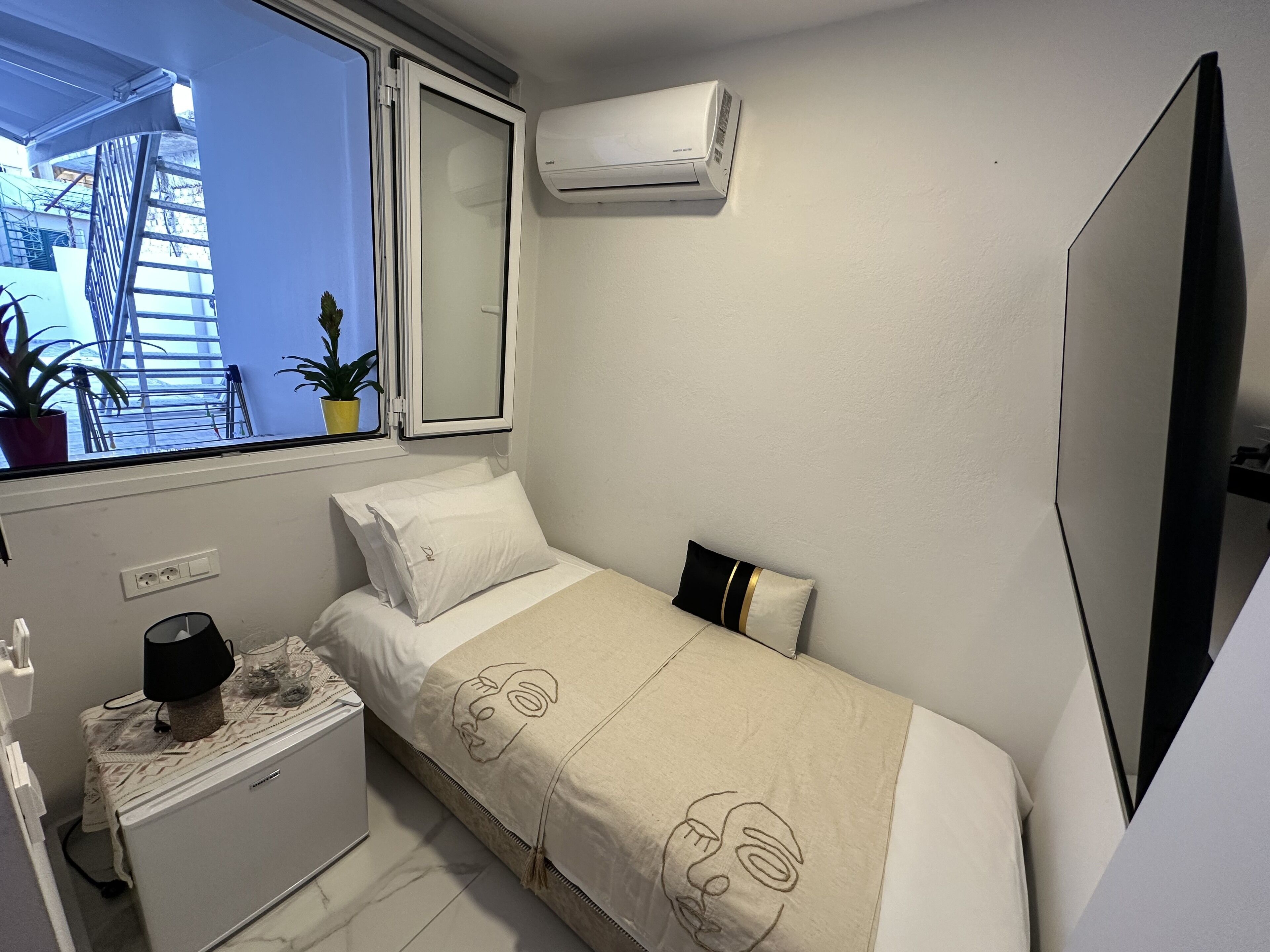 Un dormitorio pequeño y ordenado con una cama individual, una mesita de noche, aire acondicionado, una ventana con vista a edificios y una planta en maceta.