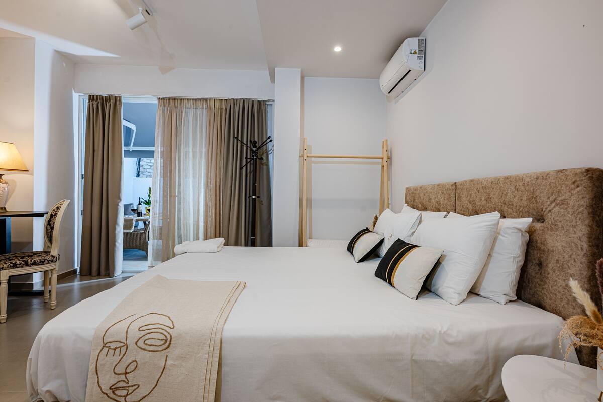 Quarto de hotel com uma cama grande, cabeceira de madeira, mesa de cabeceira, abajur e vista para o exterior através de uma janela com cortinas.