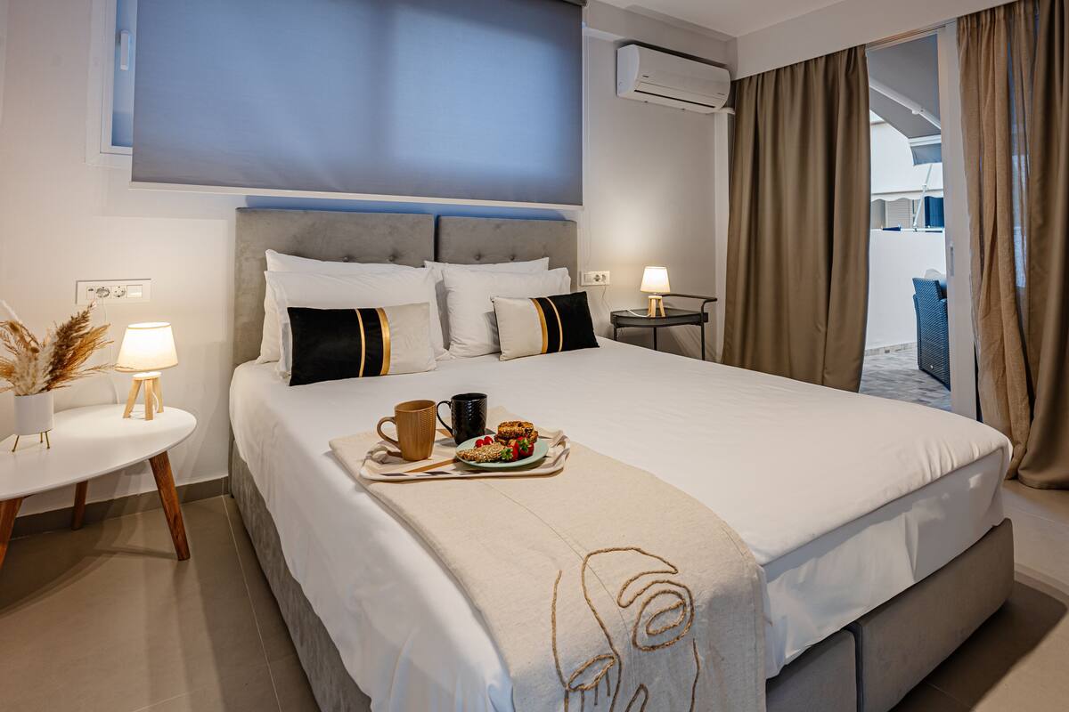 Quarto de hotel com uma cama grande, uma mesa de cabeceira com abajur e um tabuleiro com itens de café da manhã.