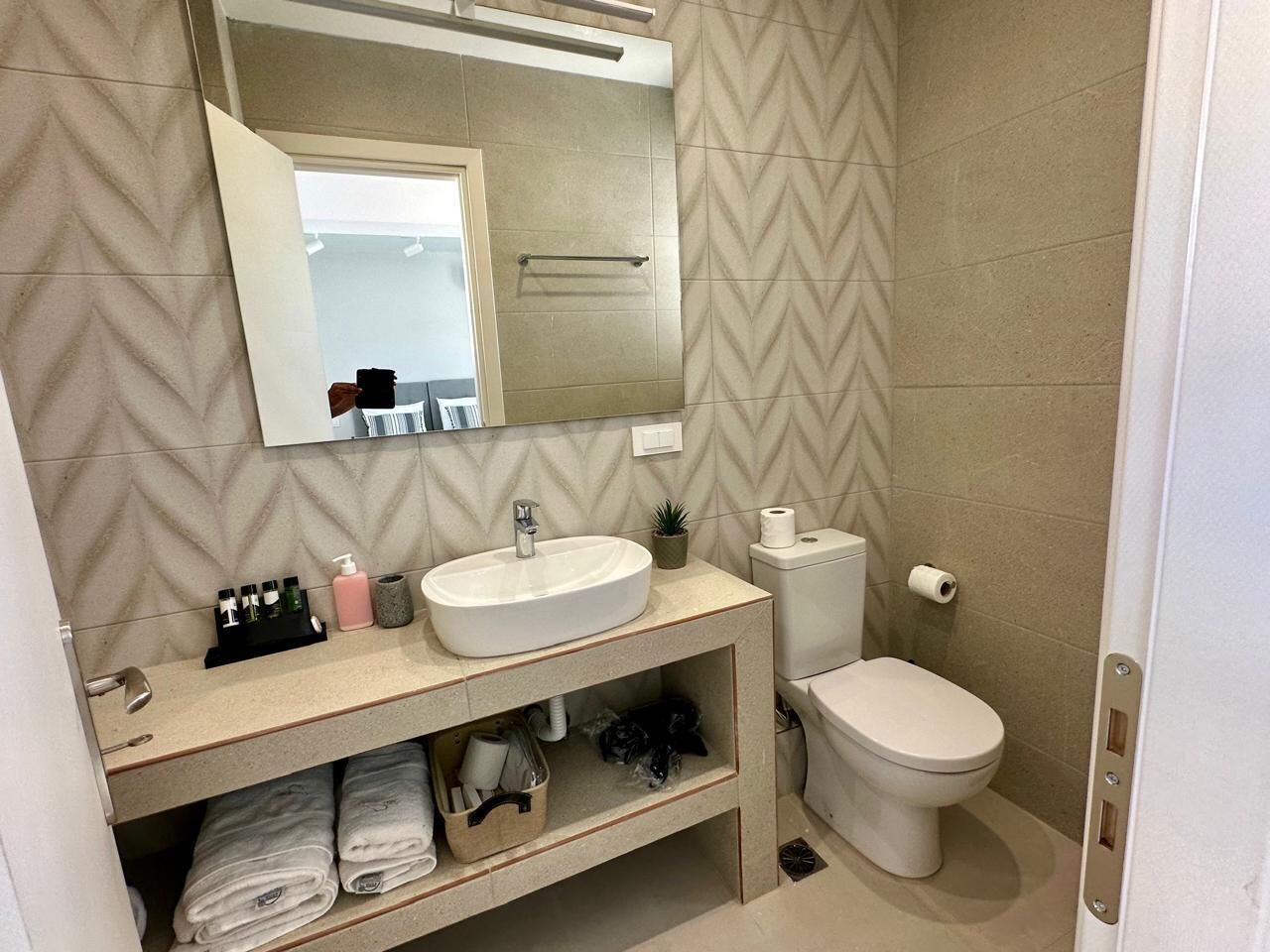 Un baño con lavamanos blanco, espejo y retrete. Hay toallas y artículos de aseo en la encimera.