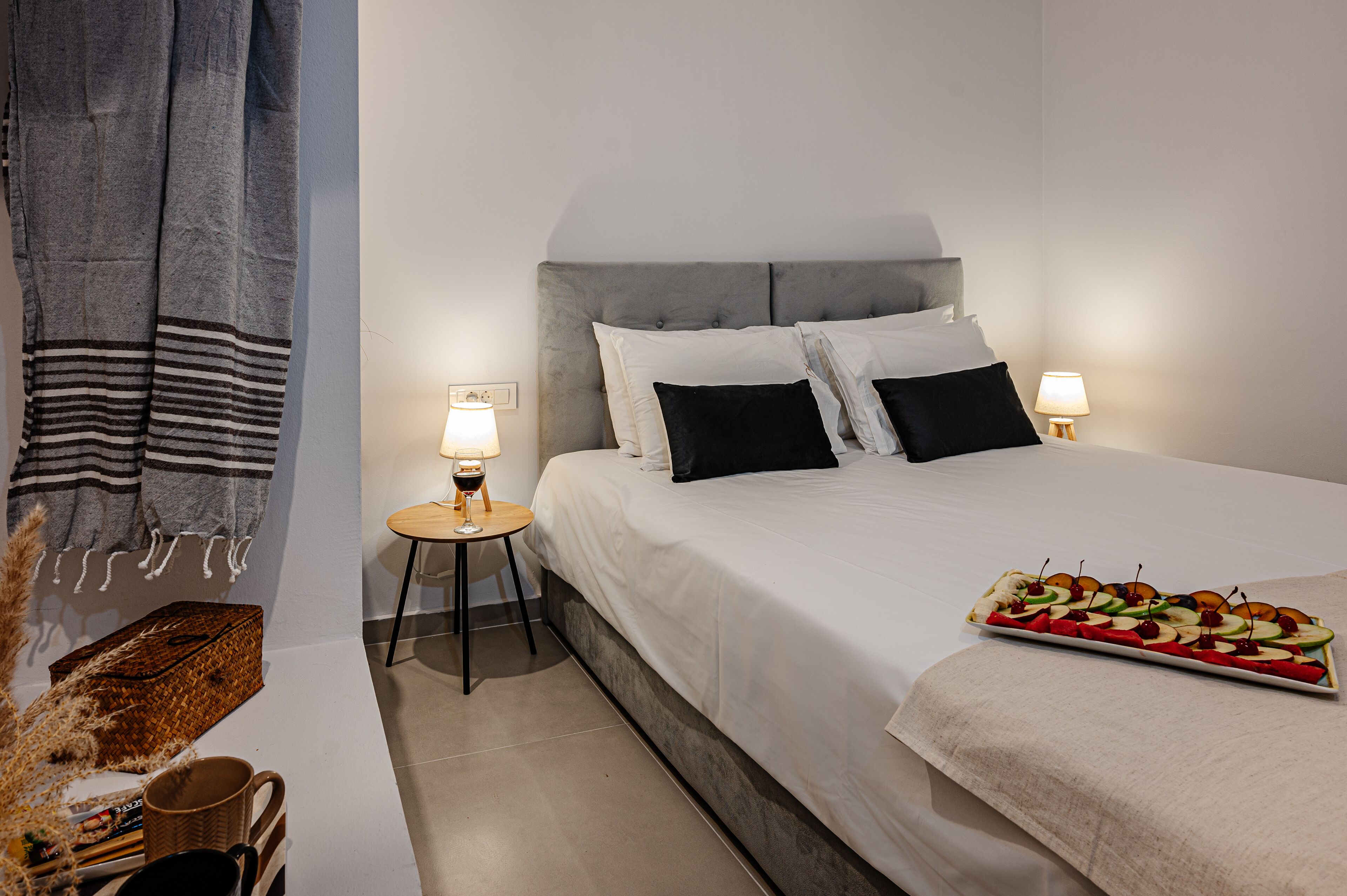 A neatly made bed with a headboard, two bedside lamps, a tray of fruit, and a striped towel on the floor.