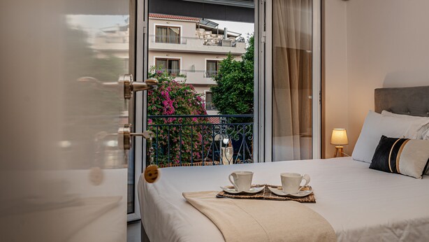 A hotel room with a bed, a balcony with a view of buildings and trees, a small table with cups and saucers, and a nightstand with a lamp.