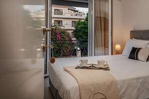 A hotel room with a bed, a balcony with a view of buildings and trees, a small table with cups and saucers, and a nightstand with a lamp.