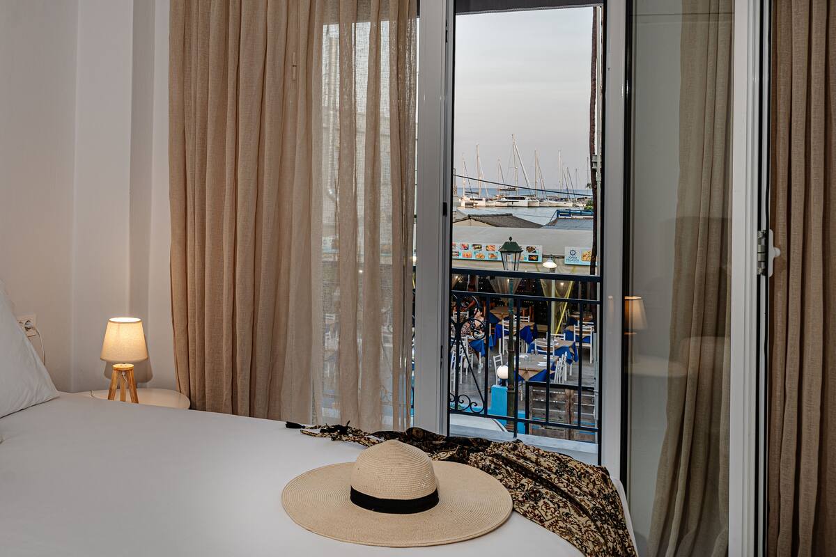 Quarto de hotel com cama, abajur, chapéu e vista para uma marina através de uma porta de vidro.