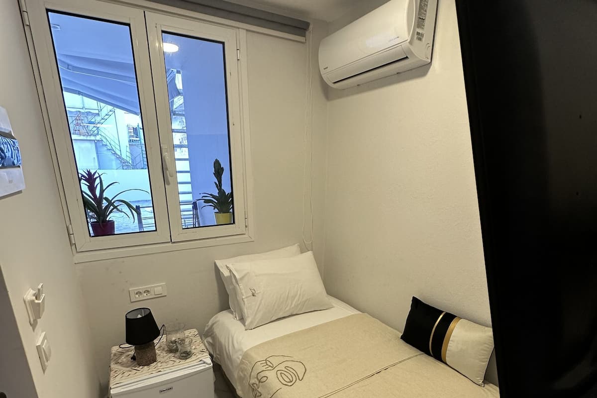 Um quarto pequeno e bem iluminado, com uma cama de solteiro, uma mesinha de cabeceira, uma janela com vista para um edifício e um ar condicionado na parede.