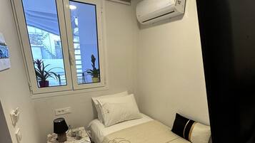 Un dormitorio pequeño y bien iluminado con una cama individual, una mesita de noche, una ventana con vistas a un edificio y un equipo de aire acondicionado en la pared.
