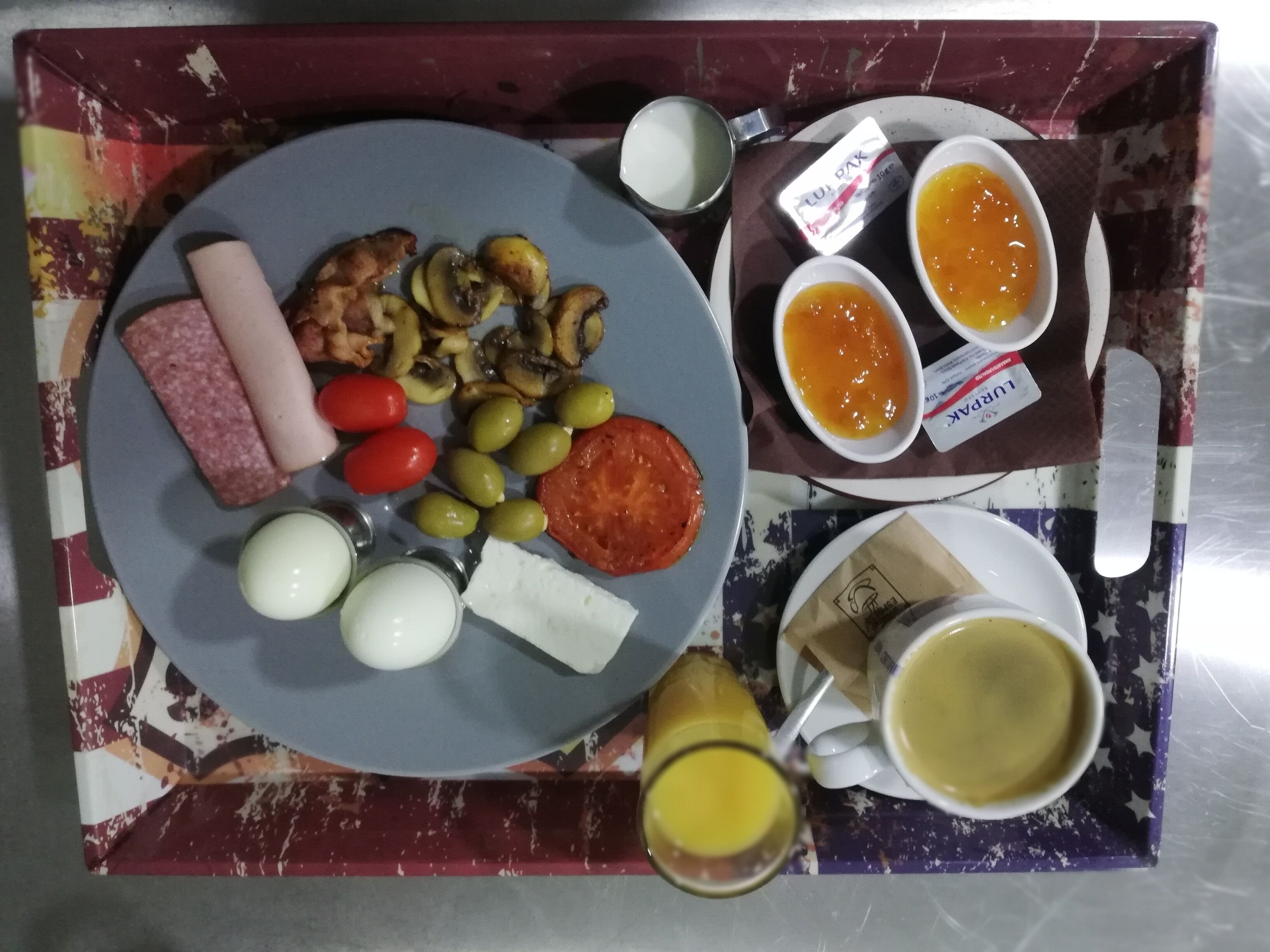 A tray with a plate of food including eggs, olives, tomatoes, and mushrooms, accompanied by two small bowls of sauce and a glass of juice.