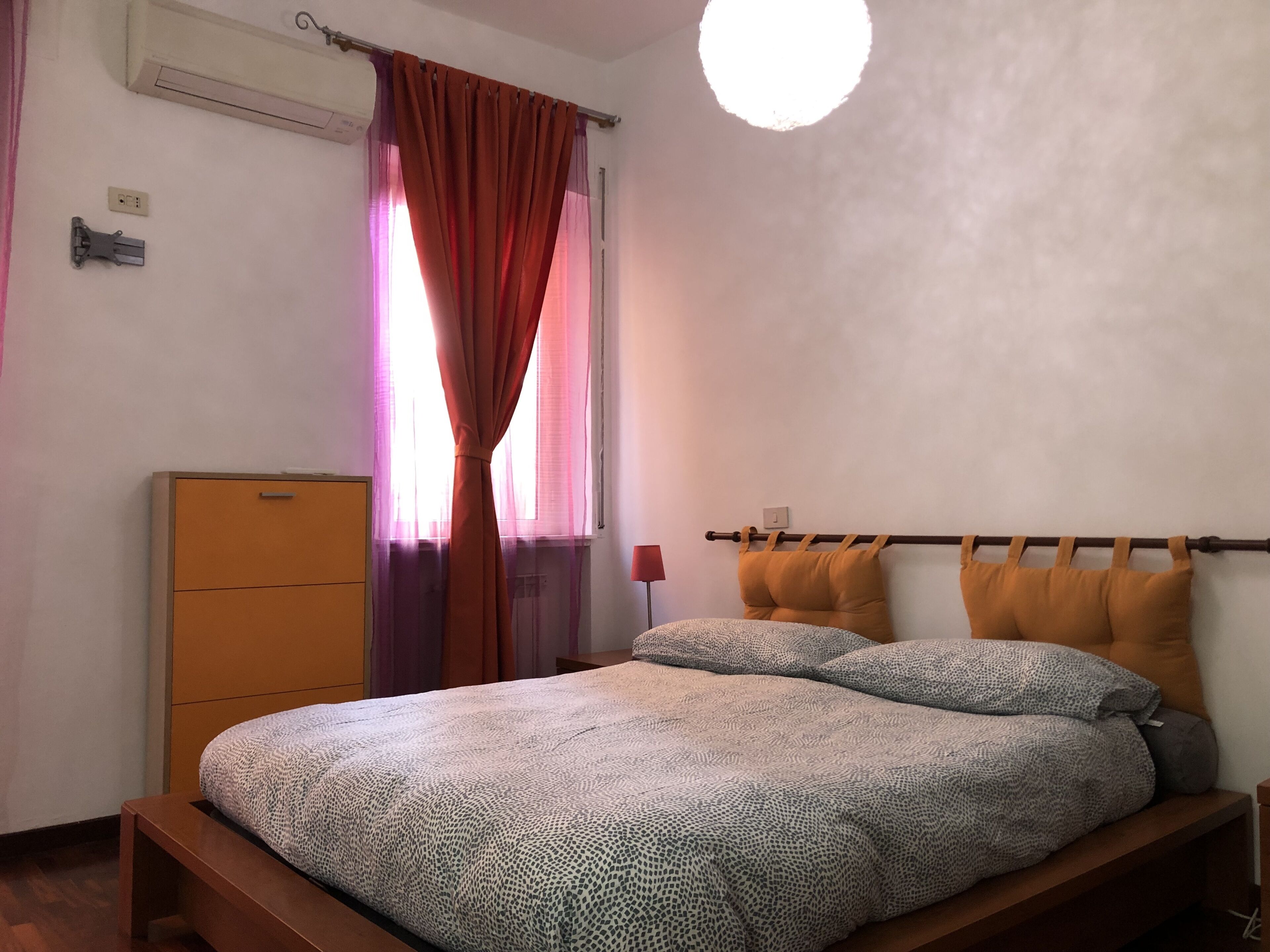 A bedroom with a bed, a wooden nightstand, a wardrobe, a window with curtains, and a ceiling light.