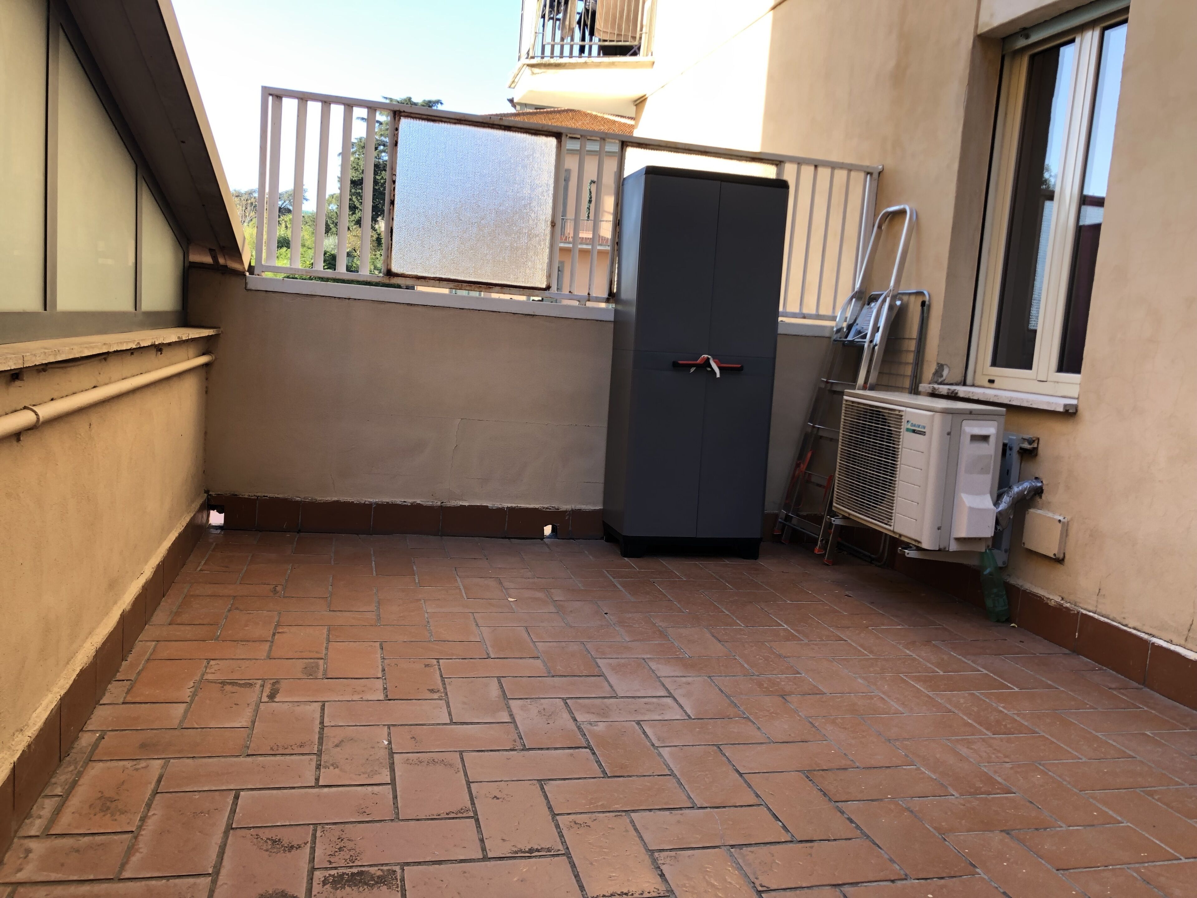 Een klein buitengebied met betegelde vloer, metalen hekwerk, een grijze kast en een airconditioning.