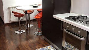 A modern kitchen with dark cabinets, a gas stove, and a white countertop. There are two red bar stools and a white table with a small dish. A colorful tile runner is on the floor, and a clock is on the wall. - Vacation Home Paradise (Viterbo)