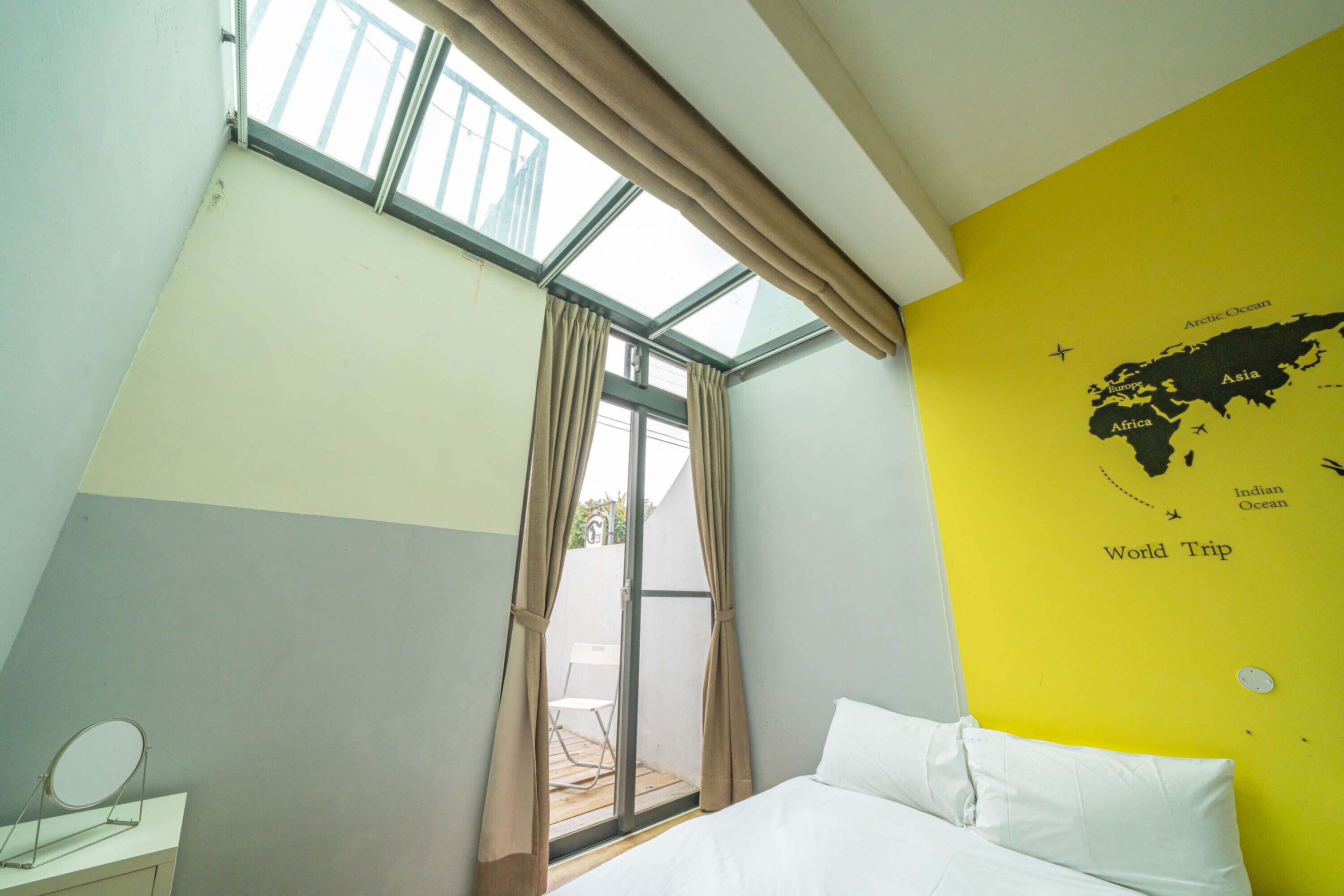 A bedroom with a bed, a window with curtains, a map on the wall, and a small table with a mirror.