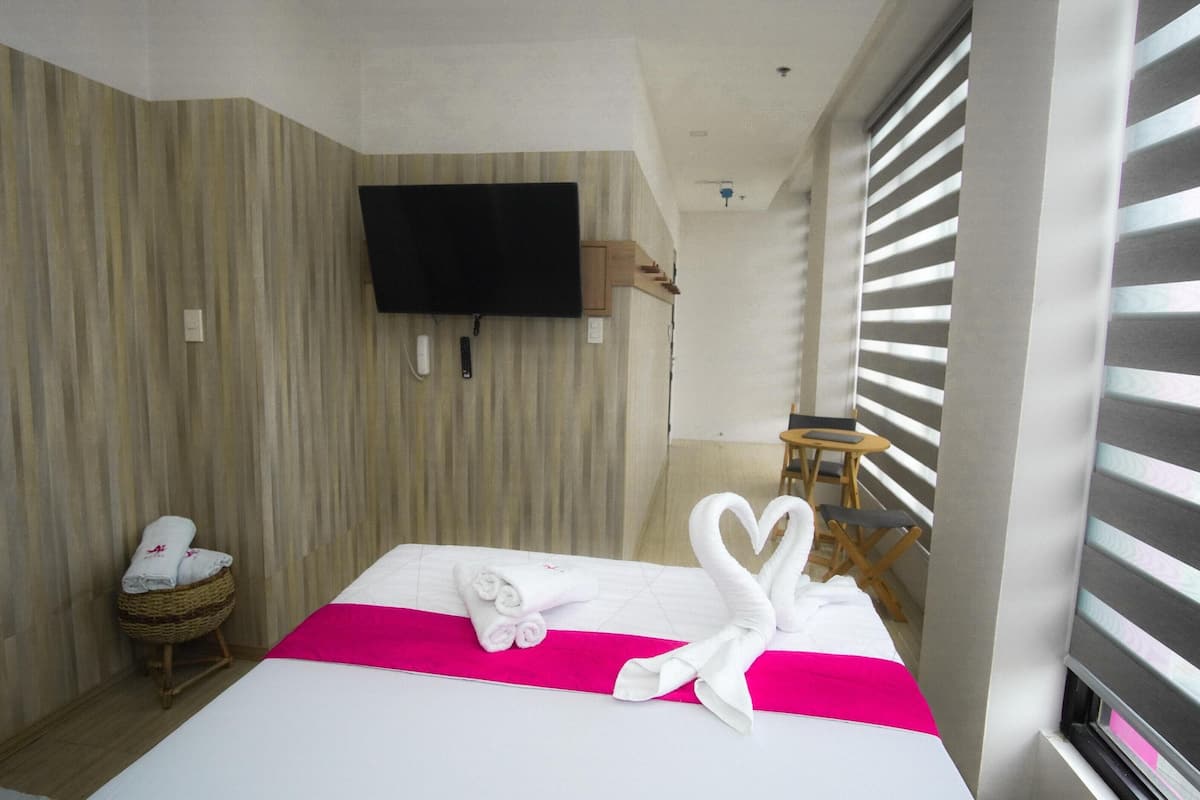 A hotel room with a bed, a television mounted on the wall, a small table, and a window with blinds.