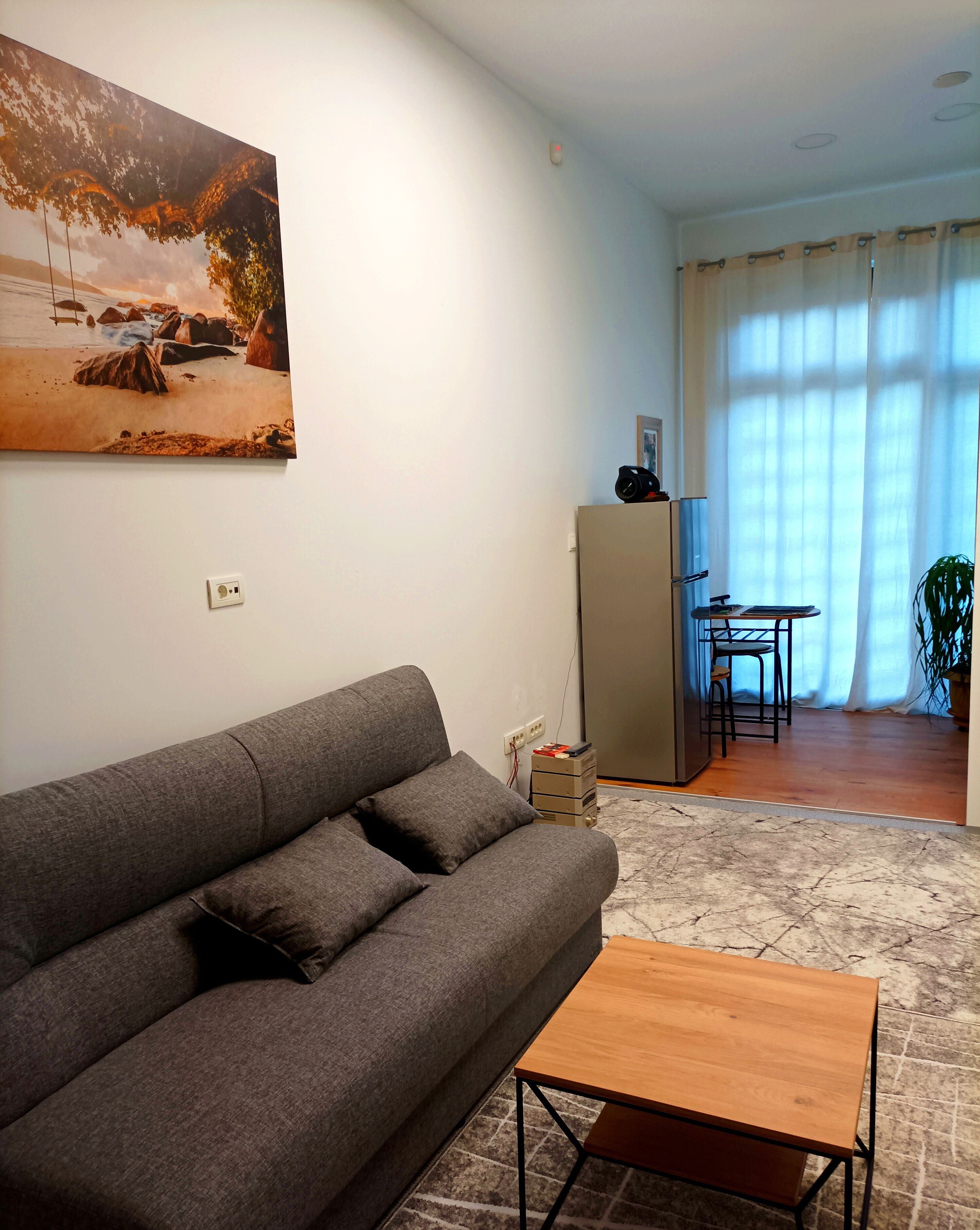 A living room with a grey sofa, a wooden coffee table, a painting of a beach scene, and a kitchen area with a fridge and a table.