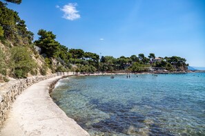 A coastal road with clear water leading to a beach, surrounded by trees and buildings.