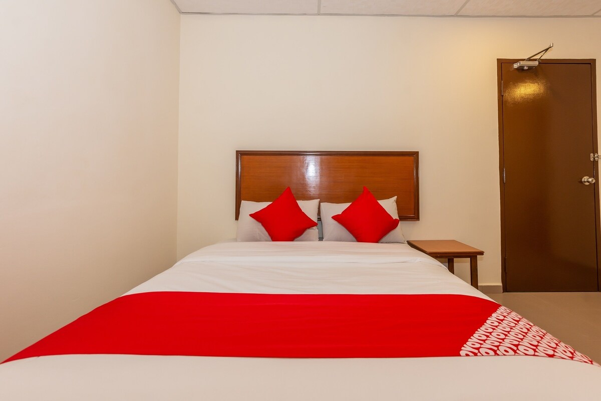 A hotel room with a bed, a wooden headboard, two red pillows, a small wooden nightstand, and a closed door.