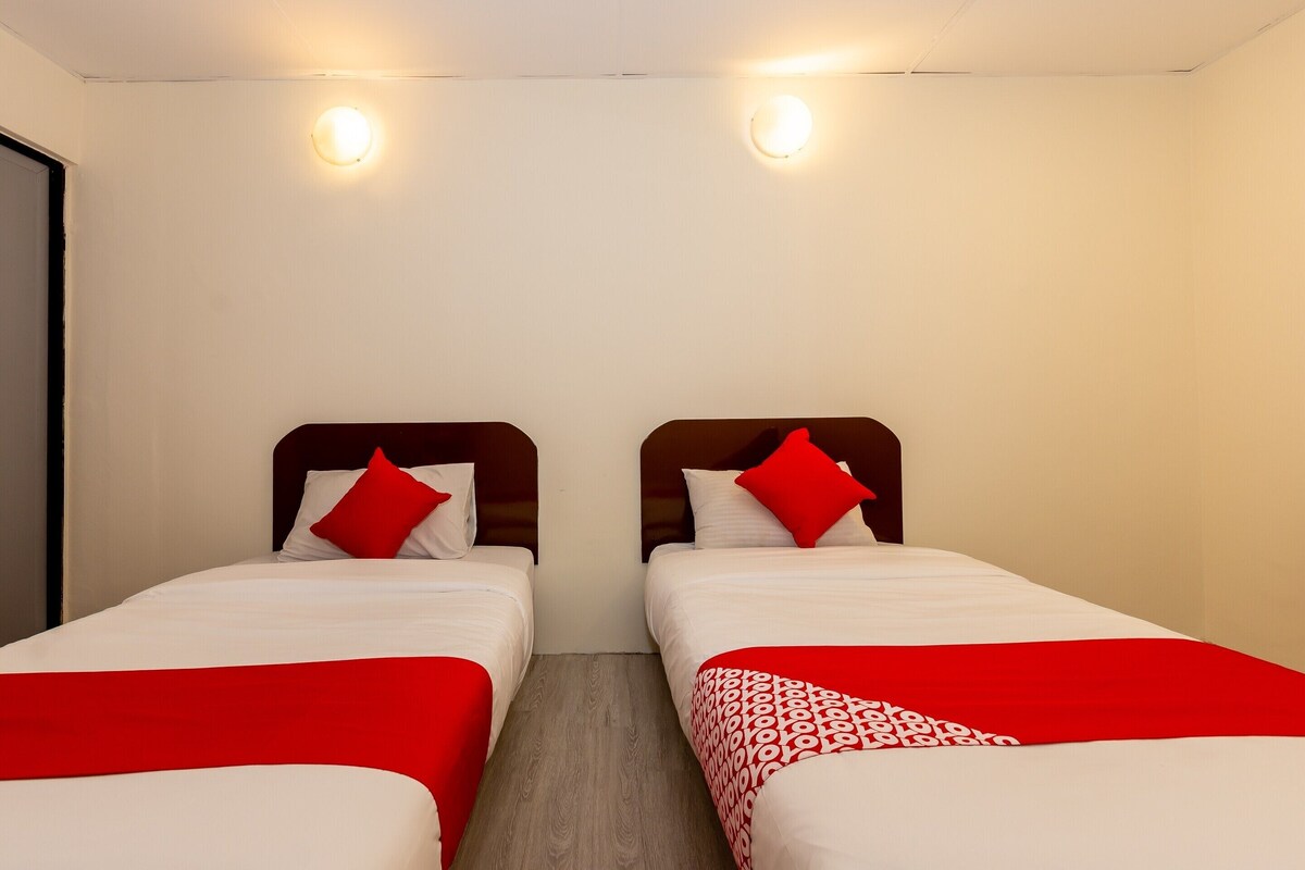 A hotel room with two single beds, each with a red and white bedspread and matching pillows. The room has a wooden floor and white walls.