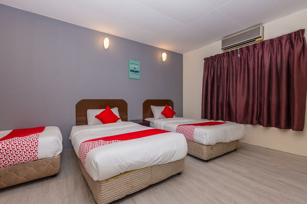 A hotel room with three single beds, each with a red and white patterned bedspread and a red pillow. The room has a light wooden floor, a grey wall, and a window with a red curtain. There is a sign on the wall and an air conditioning unit above the window.