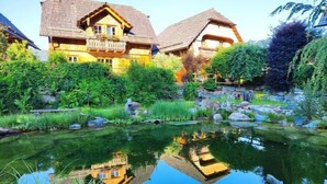 A traditional chalet with a pond, surrounded by lush greenery and a stone wall.