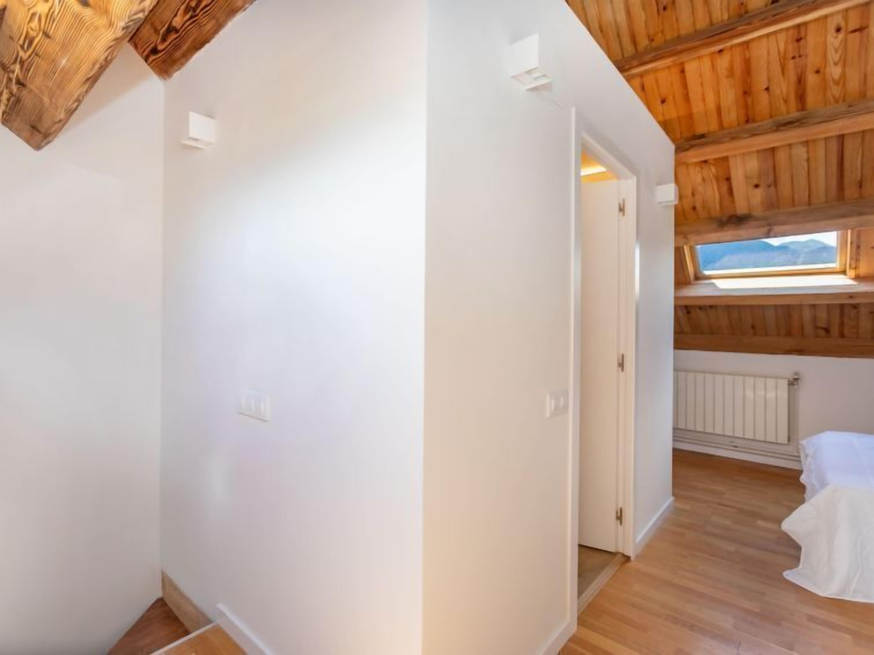 A room with white walls, wooden ceiling beams, a wooden floor, a window with a view, and a radiator.