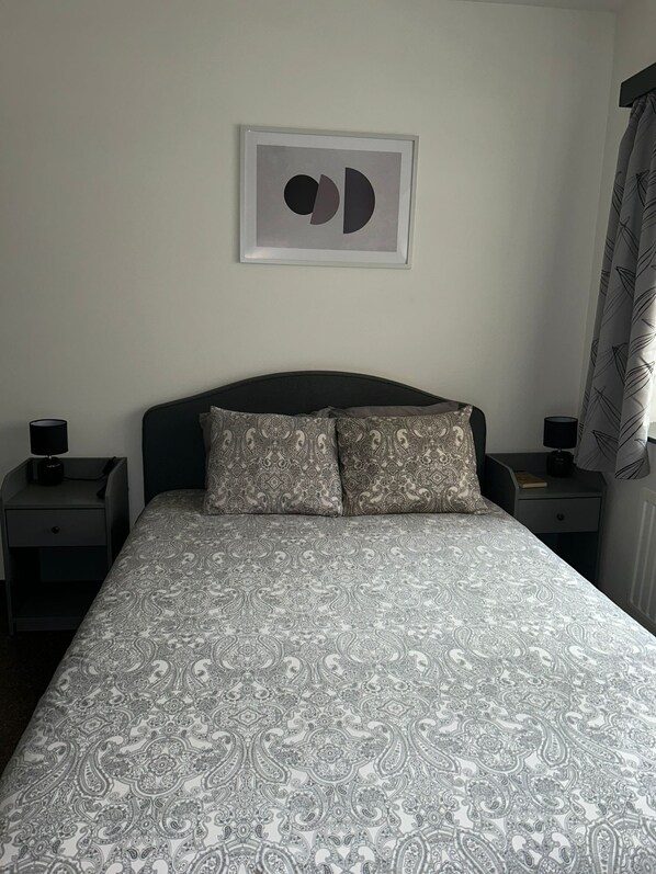 Une chambre à coucher avec un lit, deux tables de chevet, un tableau encadré au mur et une fenêtre avec des rideaux.