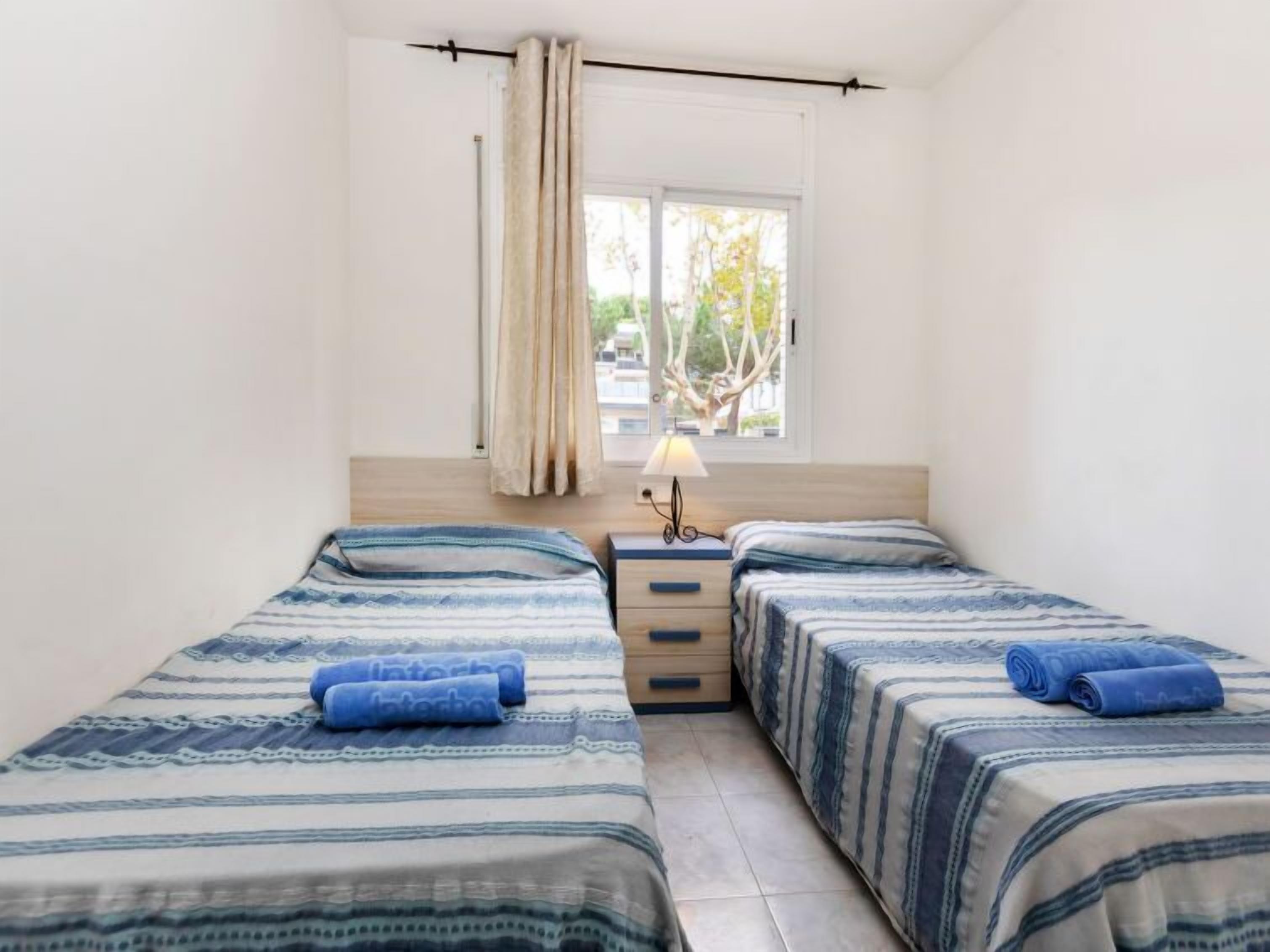A small, clean room with two beds, a window with curtains, a bedside table with a lamp, and a view of trees outside.