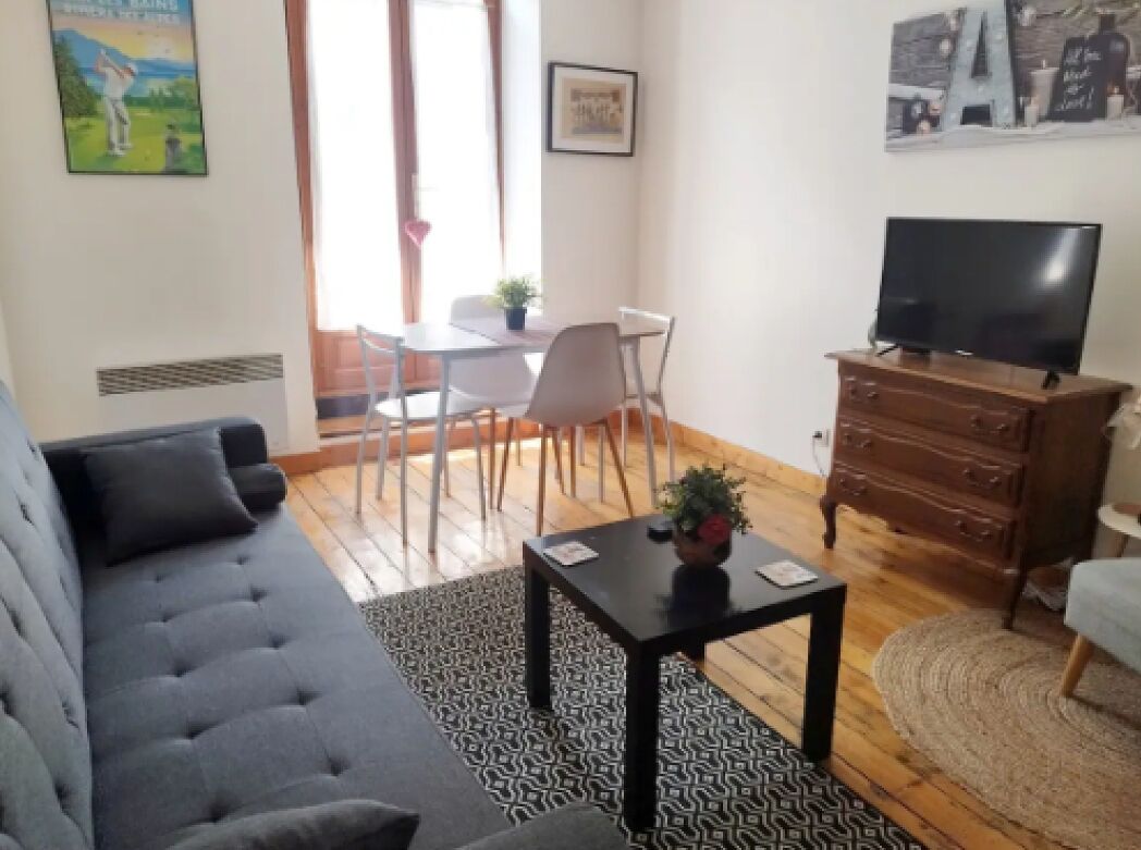 A living room with a sofa, a coffee table, a TV on a wooden cabinet, and a dining area with chairs and a table.