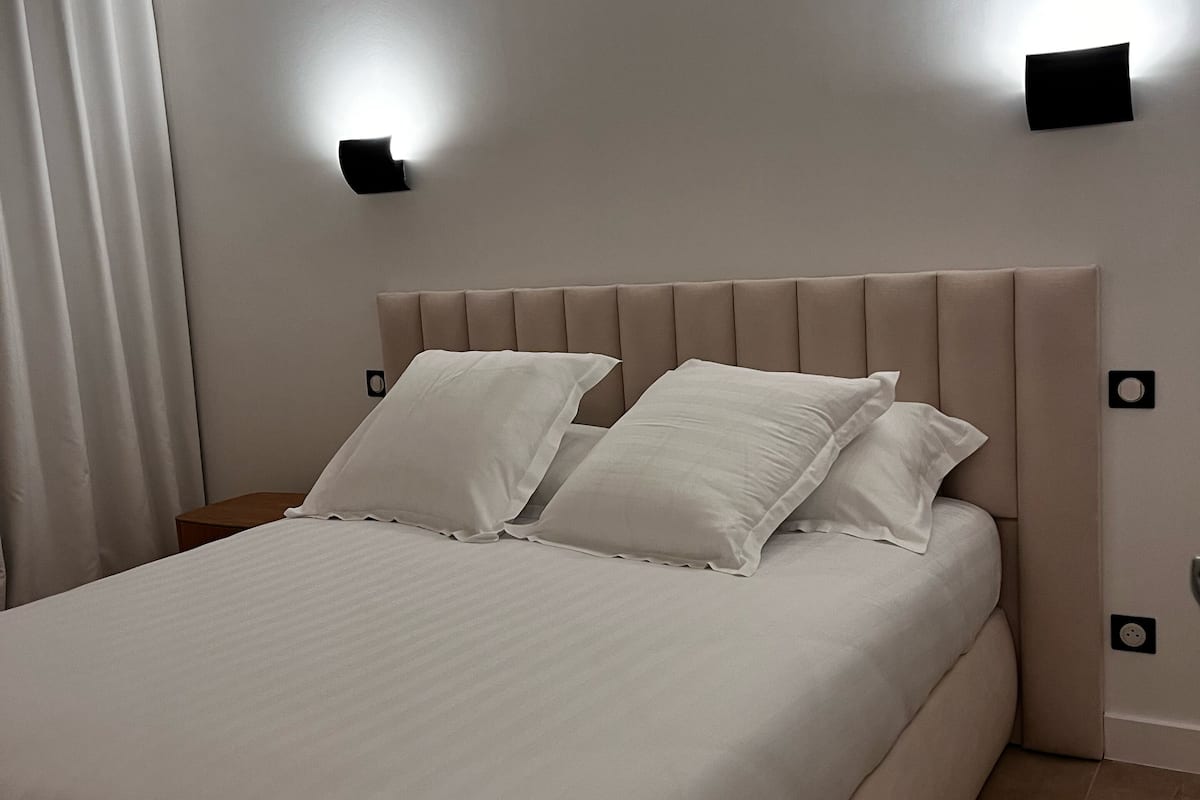 Un dormitorio moderno con una cama grande, dos lámparas fijadas a la pared y una paleta de colores neutros.