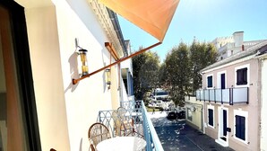 Varanda com mesa e cadeiras, com vista para uma rua com edifícios e um carro estacionado.
