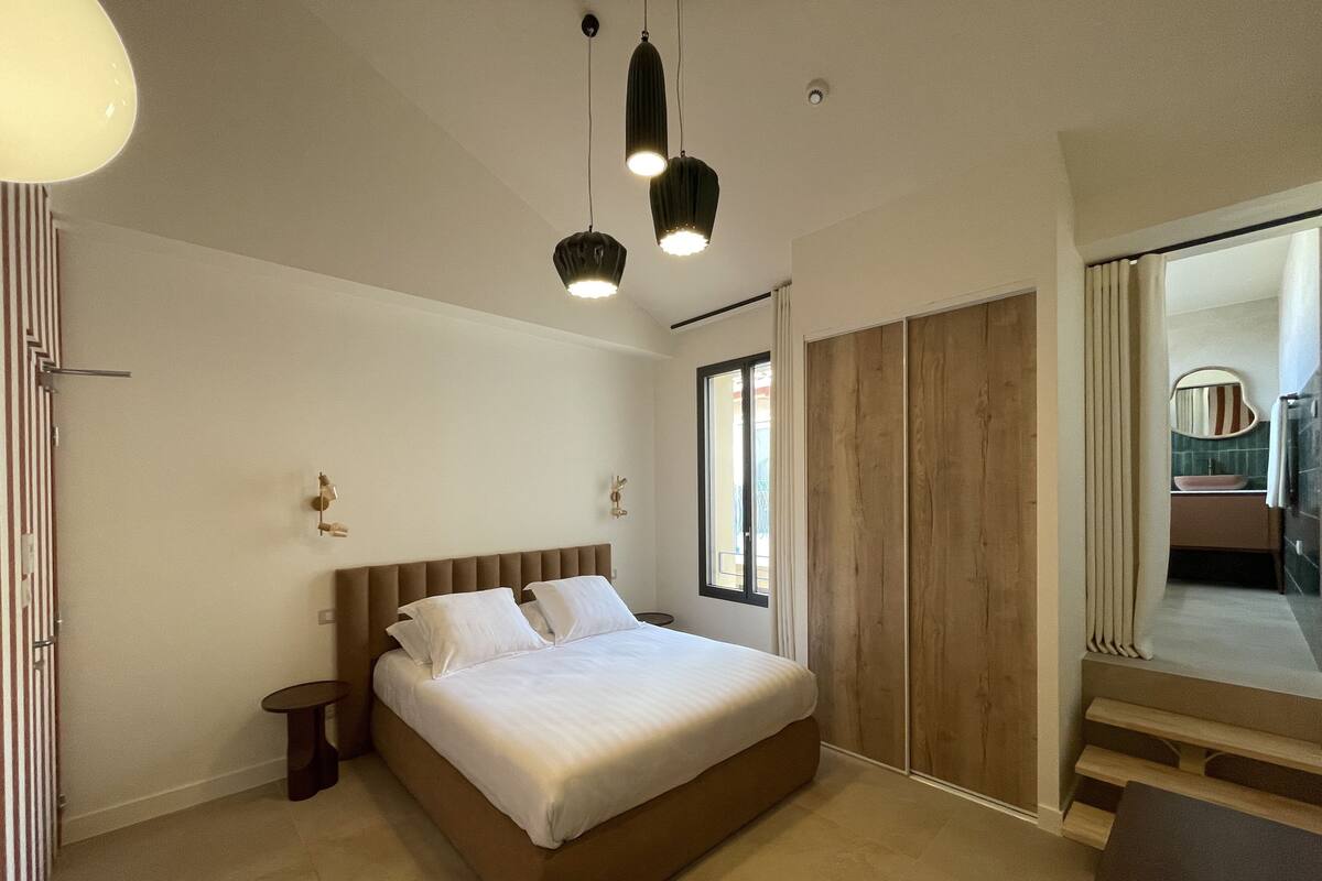 Un dormitorio con una cama grande, una mesita de noche, una pequeña mesa redonda y un espejo. Se ve una ventana con cortinas y vistas al exterior.