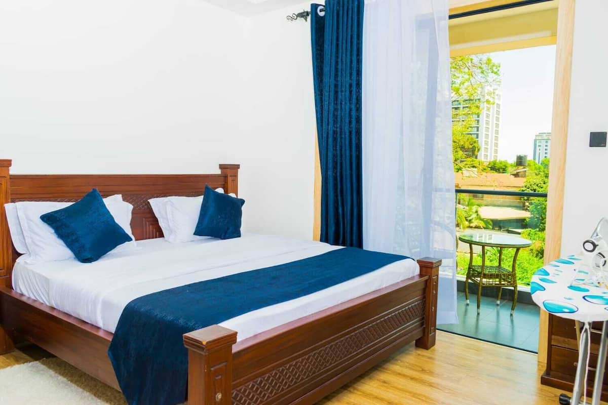 Ein Hotelzimmer mit einem hölzernen Bett, weißer Bettwäsche, einem Balkon mit Tisch und Stühlen sowie Blick auf Grünflächen und Gebäude.
