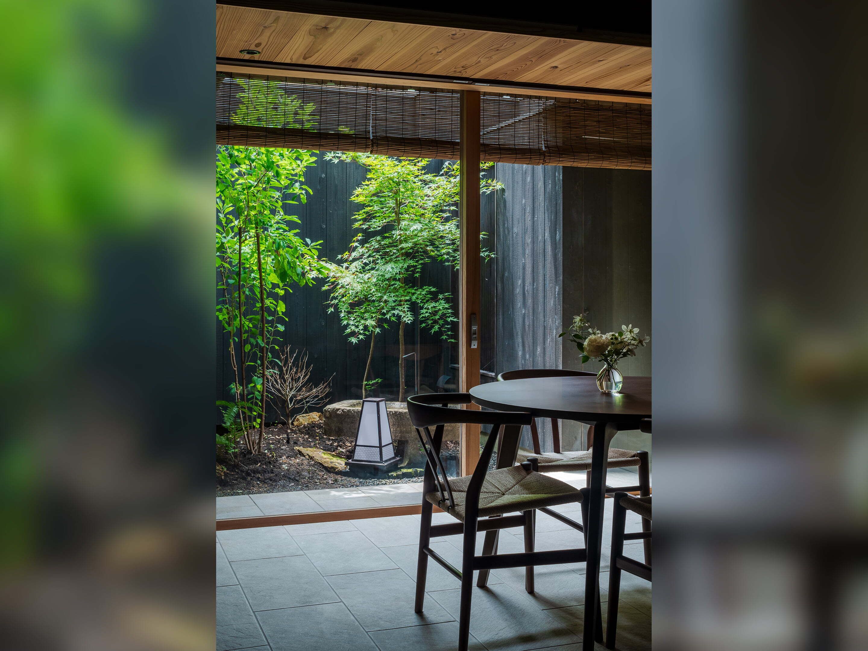 A dining area with a table and chairs, a small vase with flowers, and a view of a garden with trees and a wooden fence.